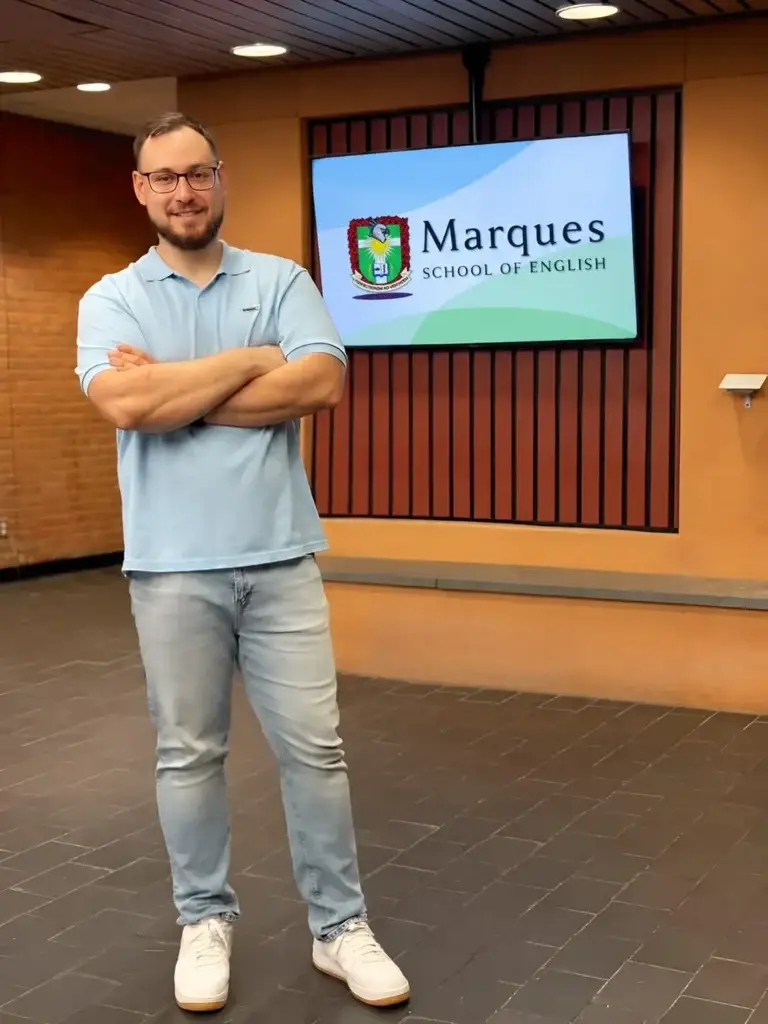 Professor da Marques School of English em pe diante do painel com a marca da escola